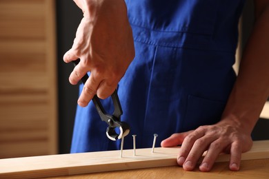 Photo of Professional repairman pulling metal nail out of wooden plank with pincers indoors, closeup