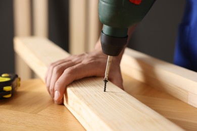 Photo of Professional repairman twisting screw into wooden plank with electric screwdriver indoors, closeup