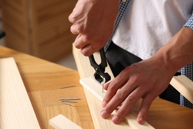 Photo of Man pulling metal nail out of wooden plank with pincers indoors, closeup