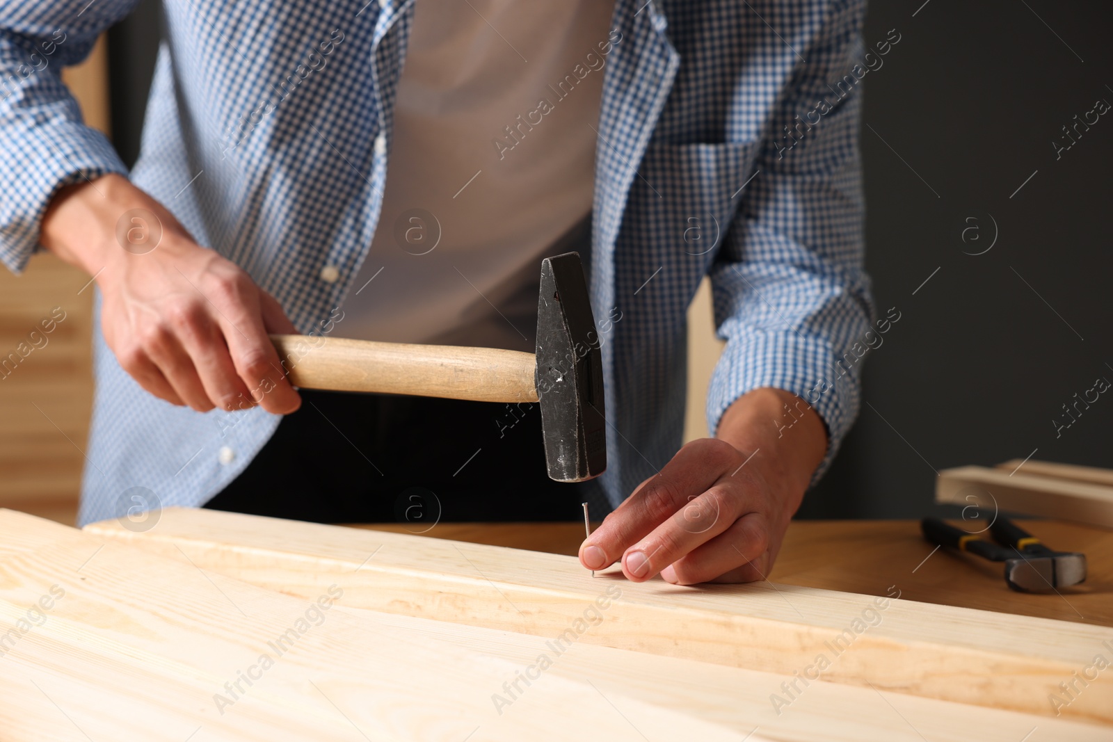 Photo of Man hammering nail into wooden plank indoors, closeup