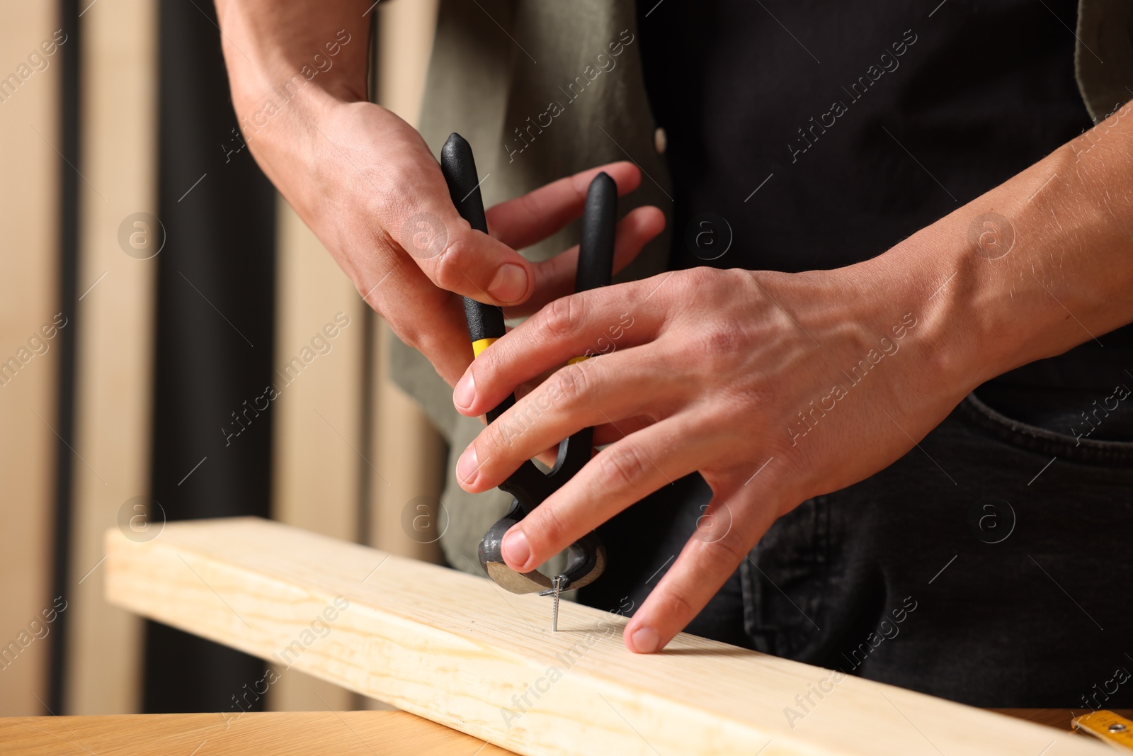Photo of Man pulling metal nail out of wooden plank with pincers indoors, closeup