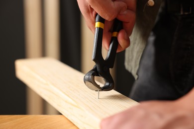 Photo of Man pulling nail out of wooden plank with pincers indoors, closeup