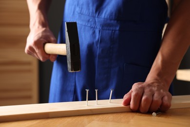 Photo of Professional repairman hammering nail into wooden plank indoors, closeup