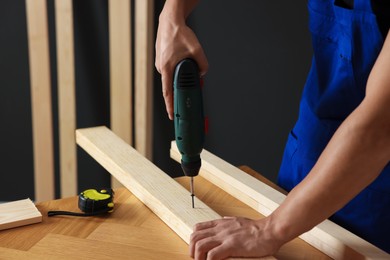 Photo of Professional repairman twisting screw into wooden plank with electric screwdriver indoors, closeup