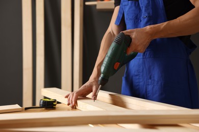Photo of Professional repairman twisting screw into wooden plank with electric screwdriver indoors, closeup