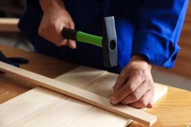 Photo of Professional repairman hammering nail into wooden plank indoors, closeup