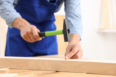 Photo of Professional repairman hammering nail into wooden plank indoors, closeup