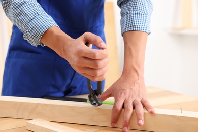 Photo of Professional repairman pulling metal nail out of wooden plank with pincers indoors, closeup