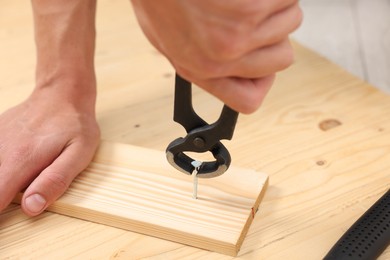 Photo of Man pulling metal nail out of wooden plank with pincers indoors, closeup