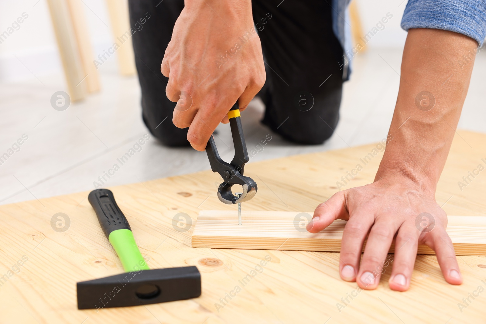 Photo of Man pulling metal nail out of wooden plank with pincers indoors, closeup