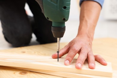 Photo of Professional repairman twisting screw into wooden plank with electric screwdriver indoors, closeup