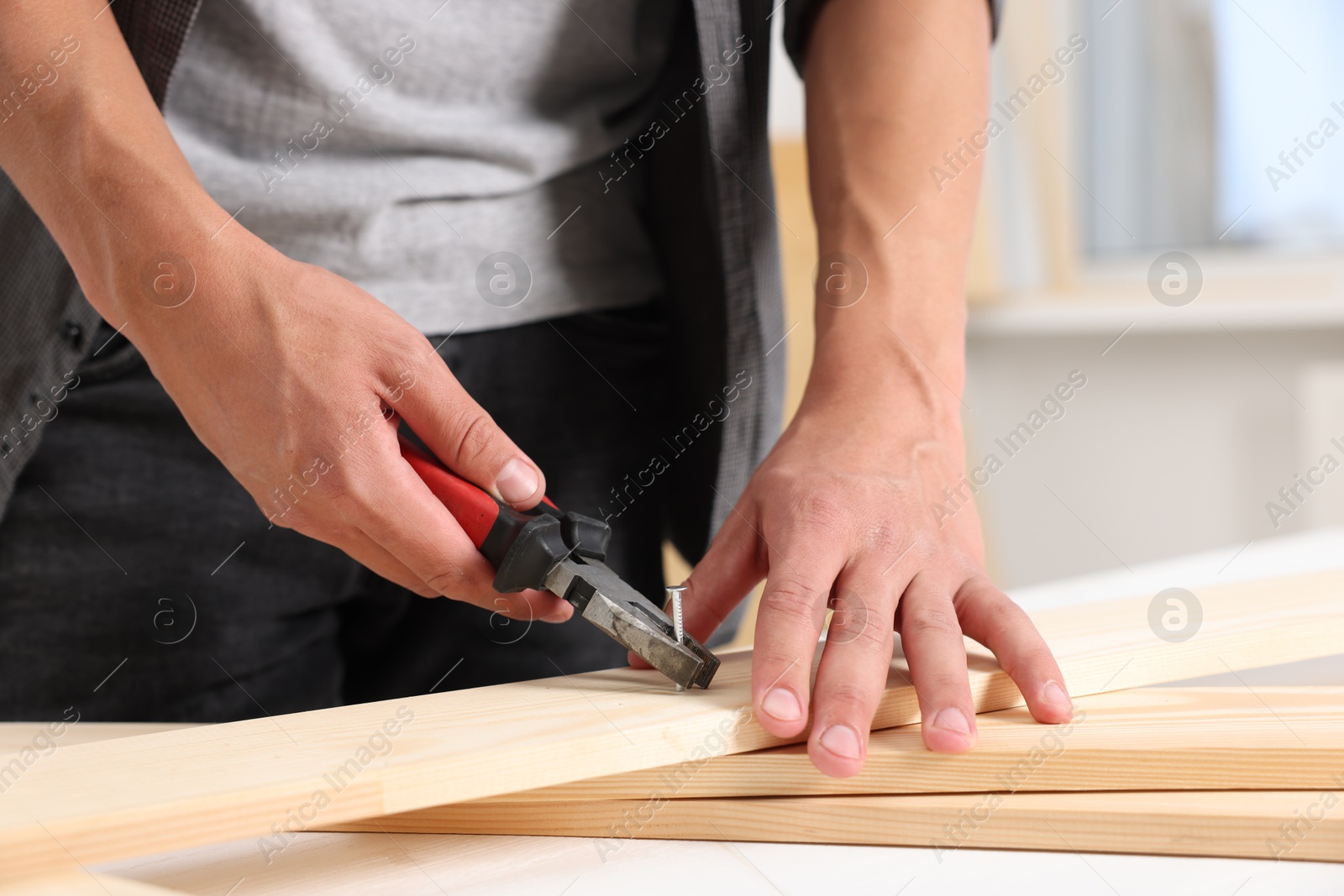 Photo of Man pulling metal nail out of wooden plank with pincers indoors, closeup