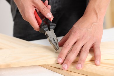 Photo of Man pulling metal nail out of wooden plank with pincers indoors, closeup