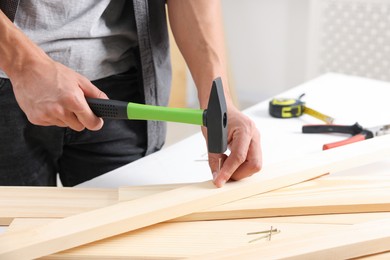 Photo of Man hammering nail into wooden plank indoors, closeup