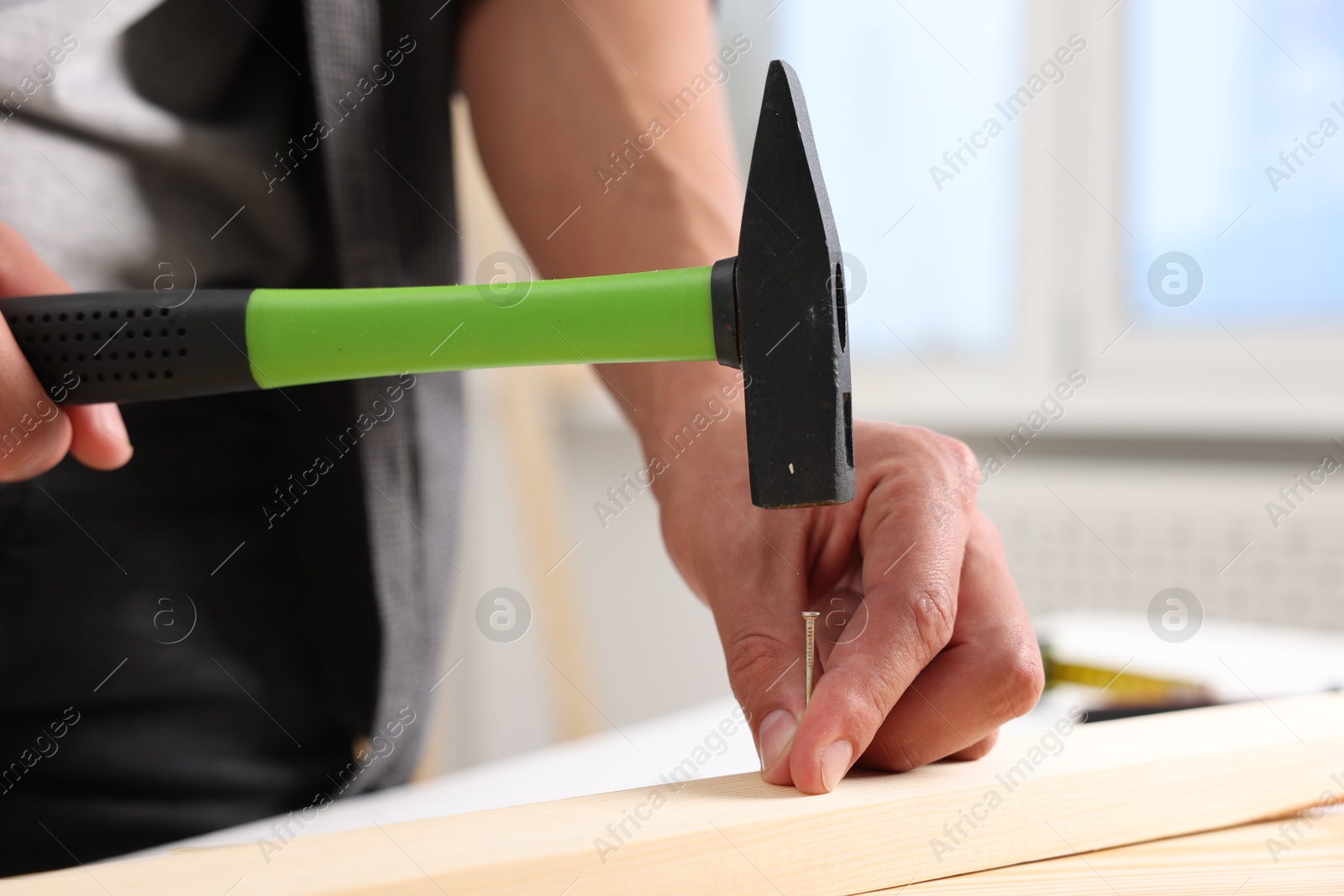 Photo of Man hammering nail into wooden plank indoors, closeup