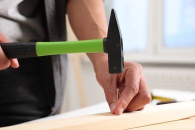 Photo of Man hammering nail into wooden plank indoors, closeup