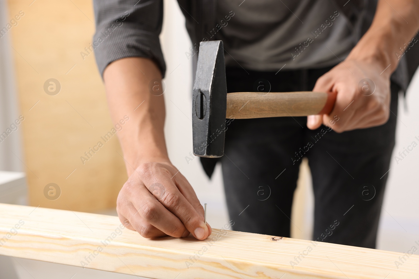 Photo of Man hammering nail into wooden plank indoors, closeup