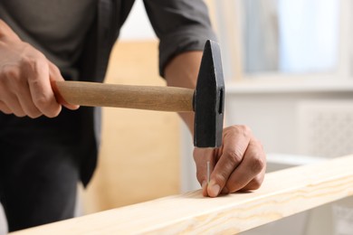 Photo of Man hammering nail into wooden plank indoors, closeup