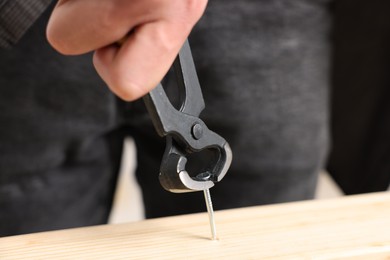 Photo of Man pulling metal nail out of wooden plank with pincers indoors, closeup