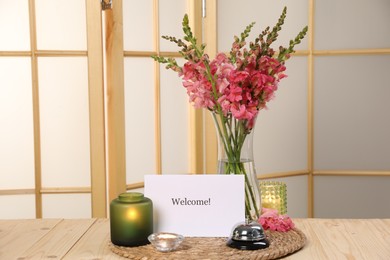 Photo of Card with word Welcome, desk bell, candles and beautiful flowers on wooden table. Space for text