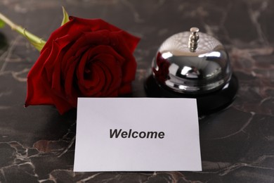 Photo of Card with word Welcome, desk bell and rose on black marble table, closeup