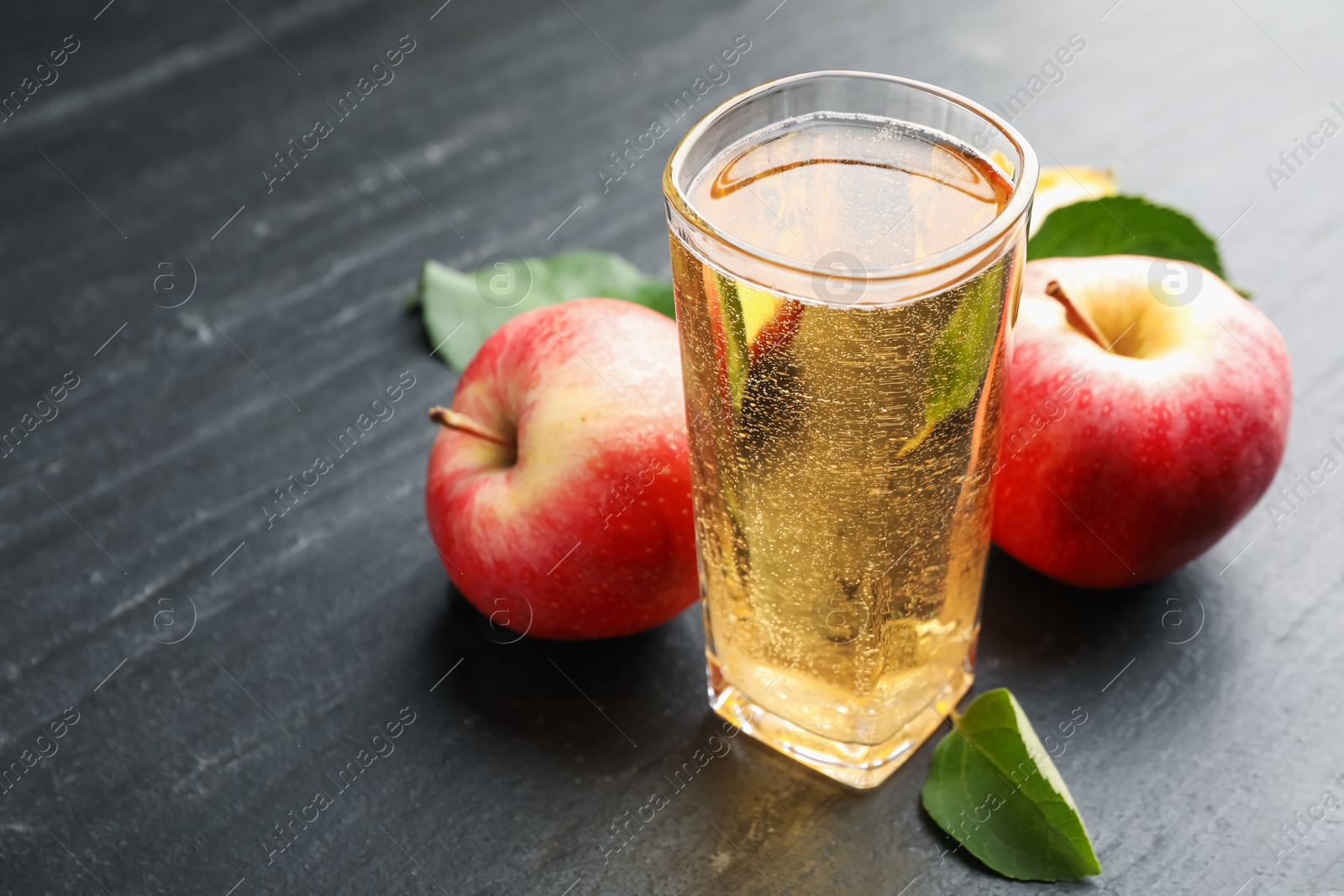 Photo of Fresh cider in glass, apples and green leaves on black table, closeup. Space for text