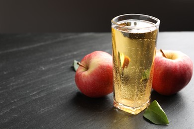 Photo of Fresh cider in glass, apples and green leaf on black table, closeup. Space for text