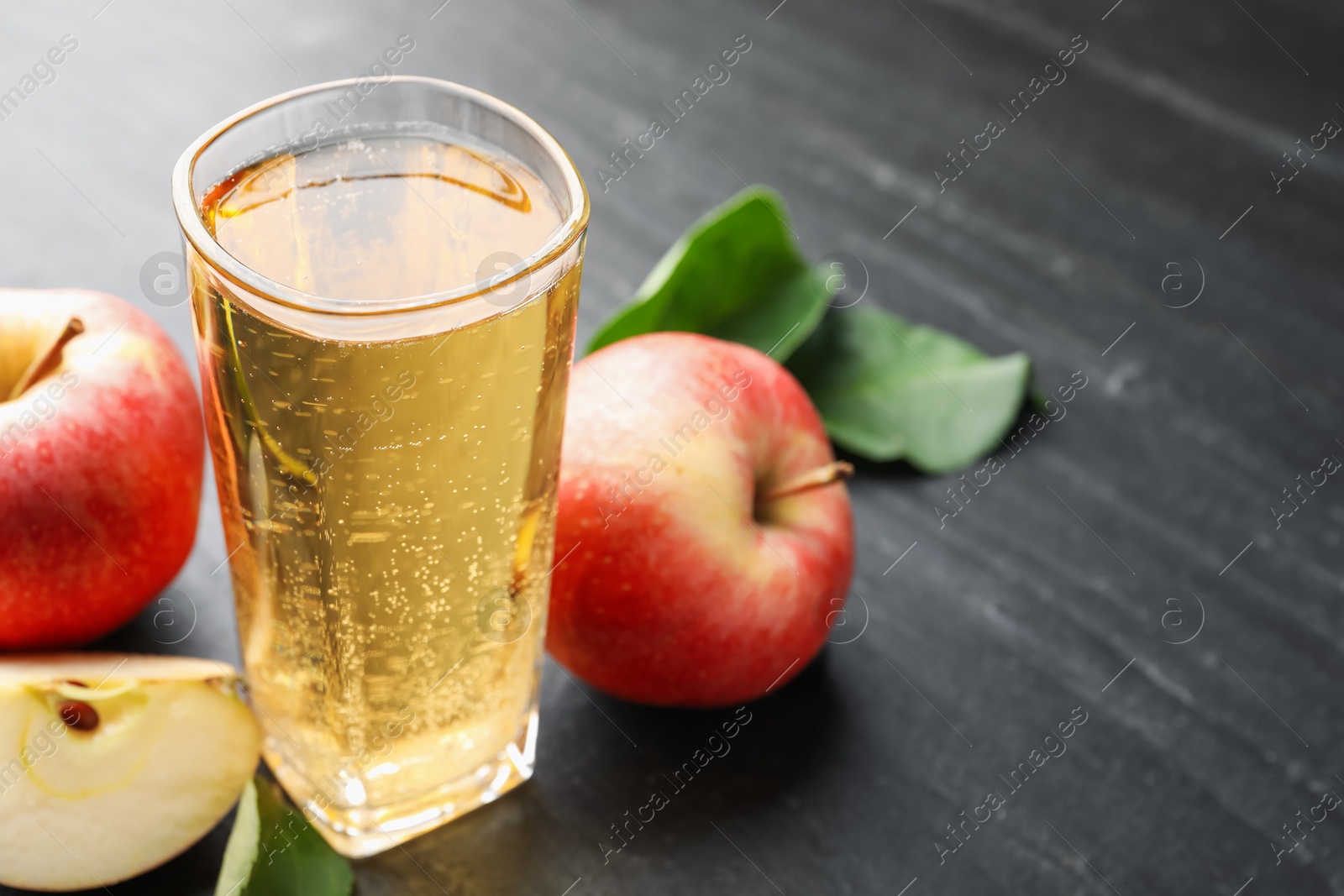 Photo of Fresh cider in glass, apples and green leaves on black table, closeup. Space for text