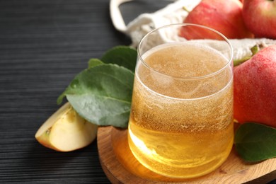 Photo of Fresh cider in glass, apples and green leaves on black textured table, closeup