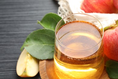 Photo of Fresh cider in glass, apples and green leaves on black textured table, closeup
