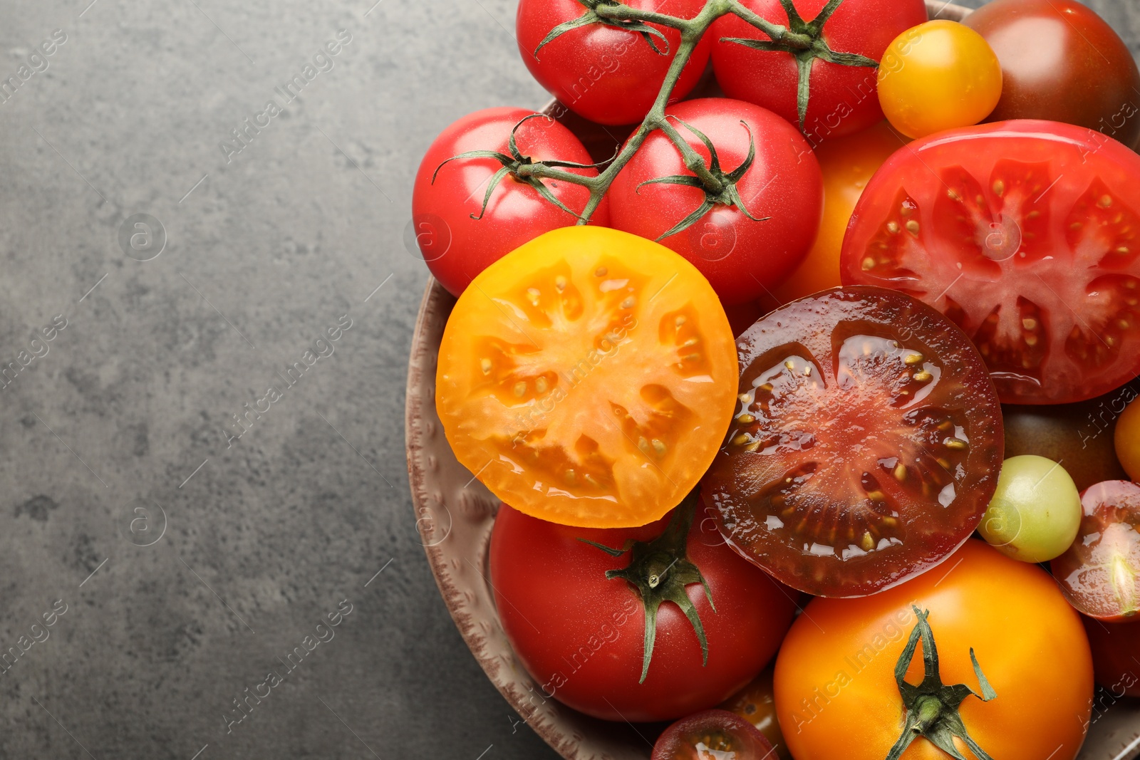 Photo of Different ripe tomatoes in bowl on grey table, top view. Space for text