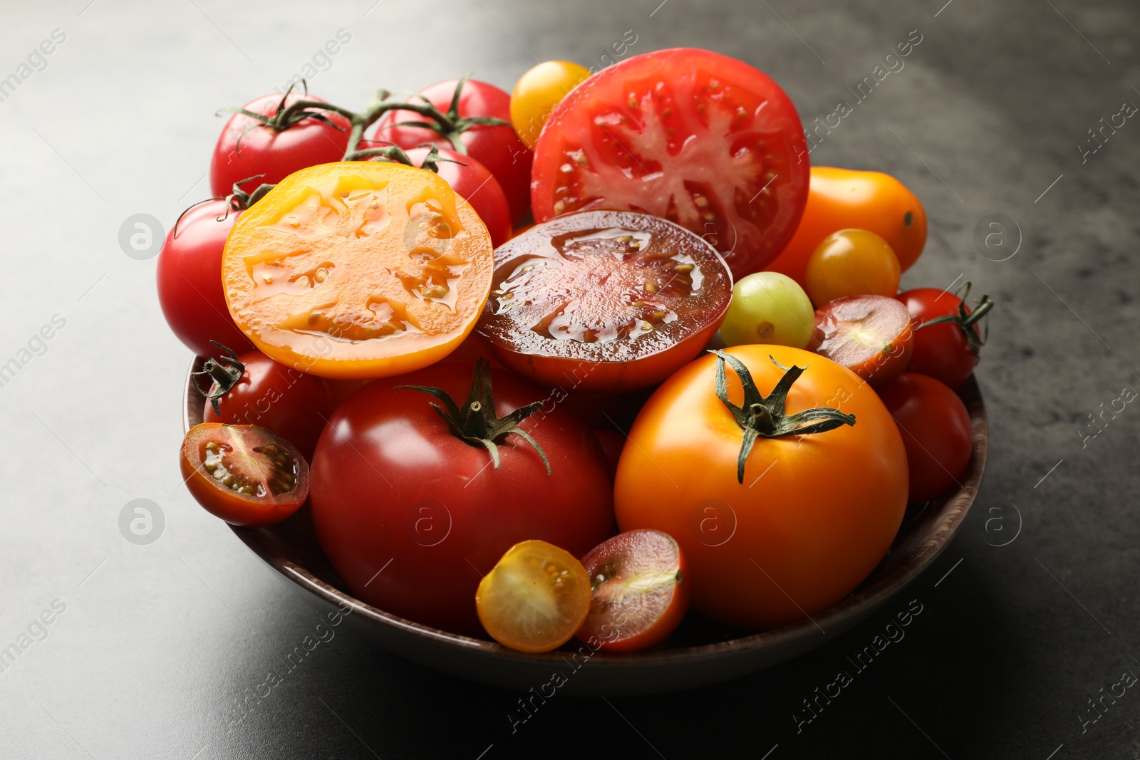 Photo of Different ripe tomatoes in bowl on grey table, closeup