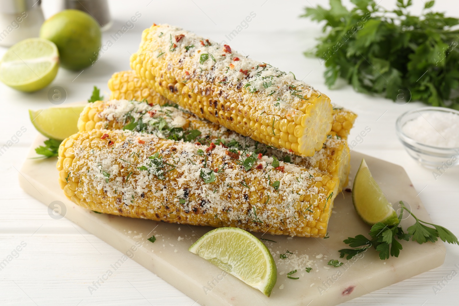 Photo of Tasty grilled corncobs with parsley, cheese and lime slices on white wooden table, closeup