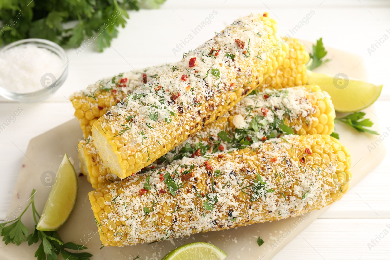Photo of Tasty grilled corncobs with parsley, cheese and lime slices on white wooden table, closeup
