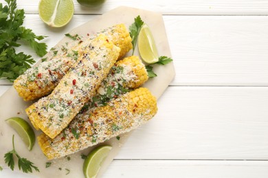 Photo of Tasty grilled corncobs with parsley, cheese and lime slices on white wooden table, flat lay. Space for text