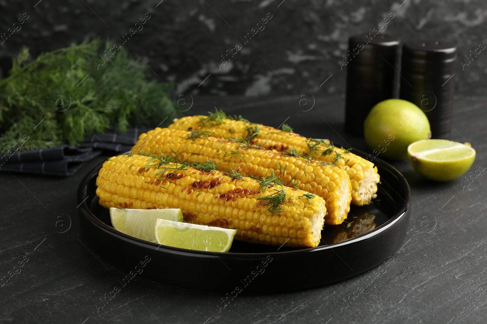 Photo of Tasty grilled corncobs with dill and lime slices on black table