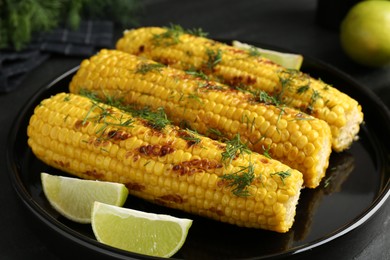 Photo of Tasty grilled corncobs with dill and lime slices on black table, closeup