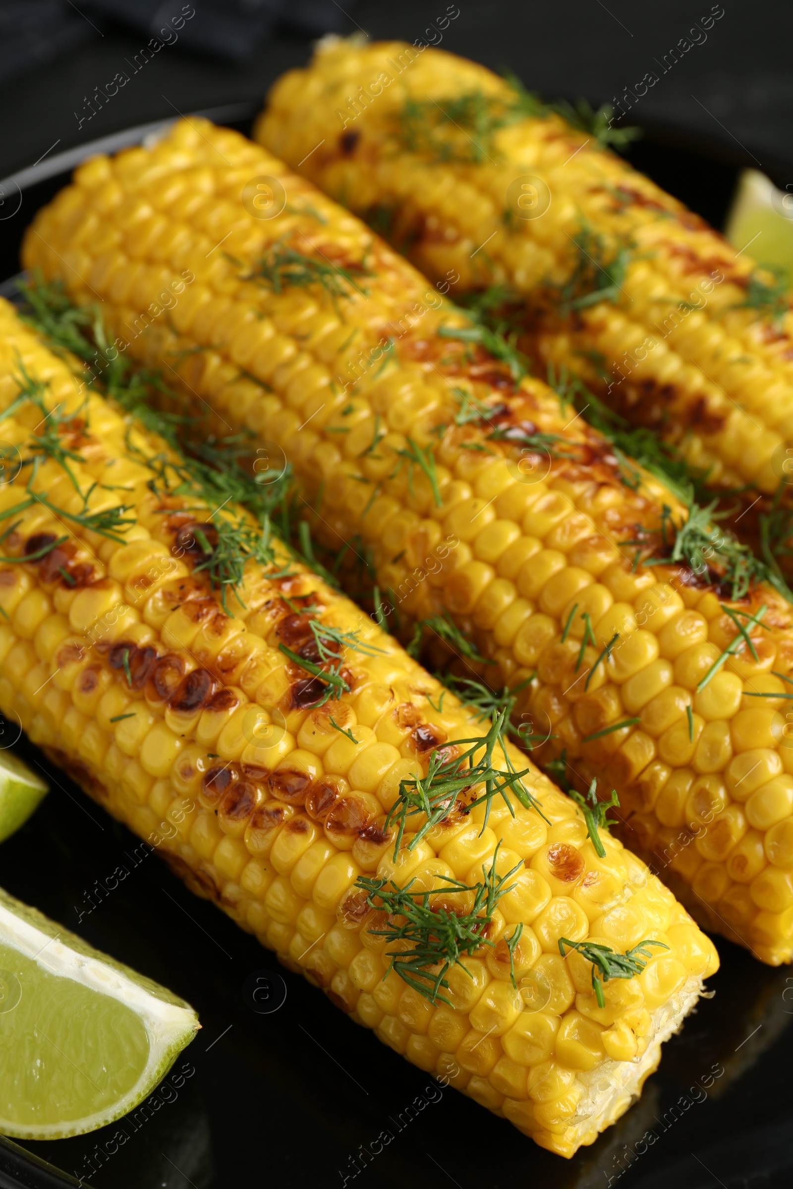 Photo of Tasty grilled corncobs with dill and lime slices on black table, closeup