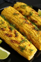 Photo of Tasty grilled corncobs with dill and lime slices on black table, closeup