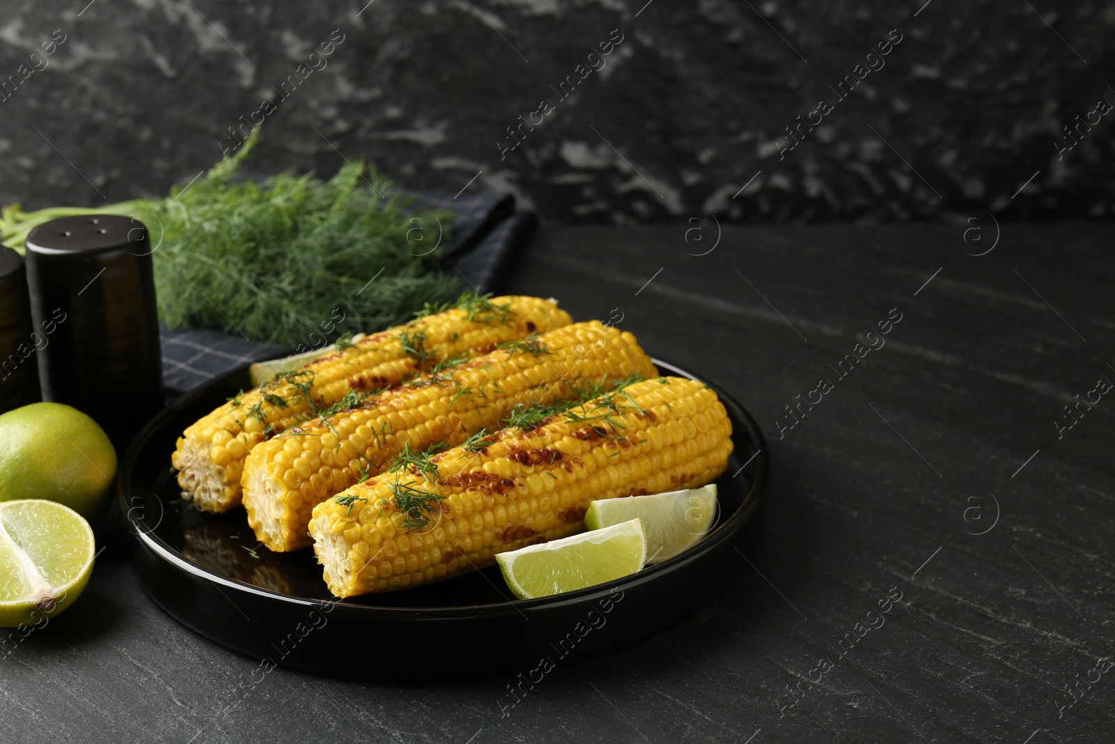 Photo of Tasty grilled corncobs with dill and lime slices on black table. Space for text