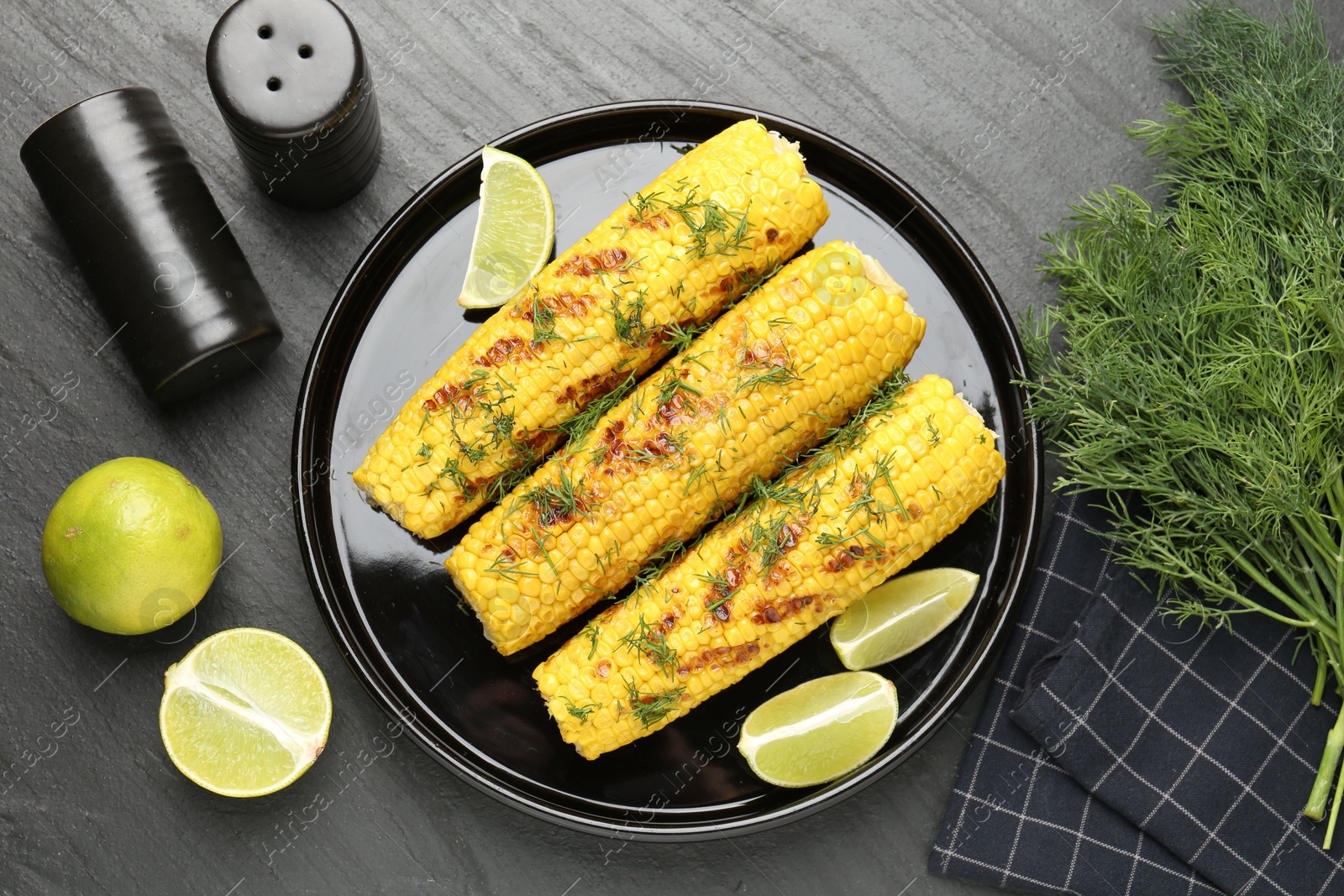 Photo of Tasty grilled corncobs with dill and lime slices on black table, flat lay