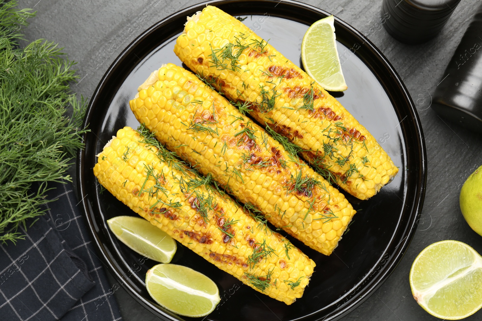 Photo of Tasty grilled corncobs with dill and lime slices on black table, flat lay