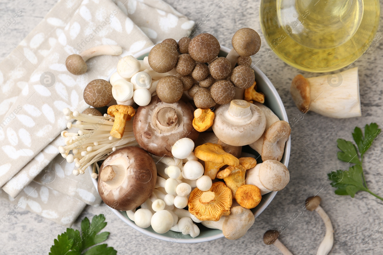 Different raw mushrooms in bowl, parsley and oil on grey textured table, flat lay Photo of Different raw mushrooms in bowl, parsley and oil on grey textured table, flat lay