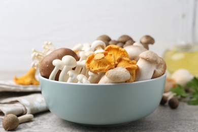 Photo of Different raw mushrooms in bowl on grey textured table against light background, closeup