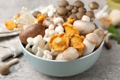 Photo of Different raw mushrooms in bowl on grey textured table, closeup