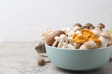 Photo of Different raw mushrooms in bowl on grey textured table against light background, closeup. Space for text