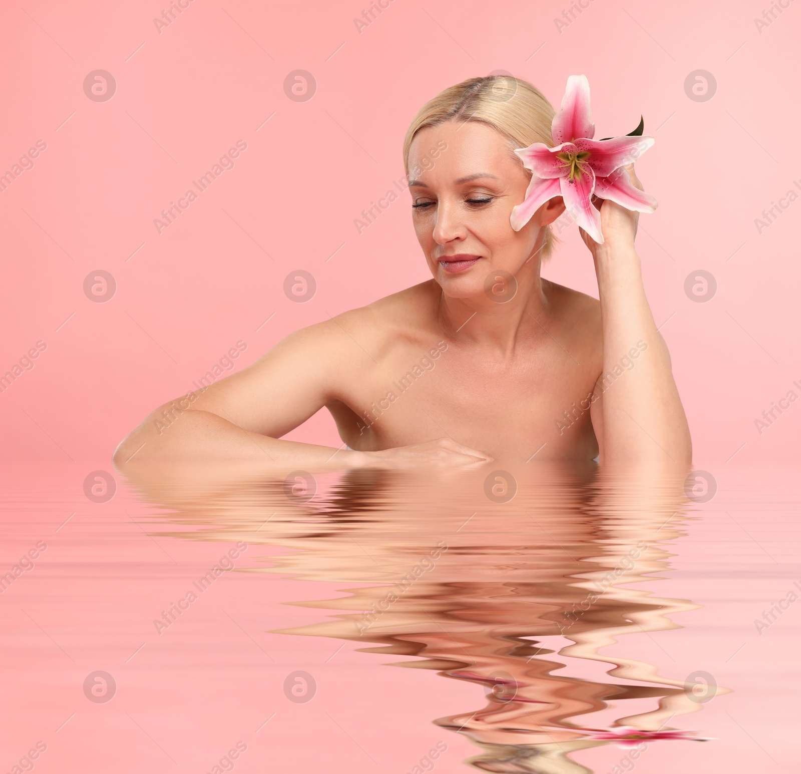 Image of Beautiful woman with perfect skin and lily flower against pink background. Her reflection in rippled water