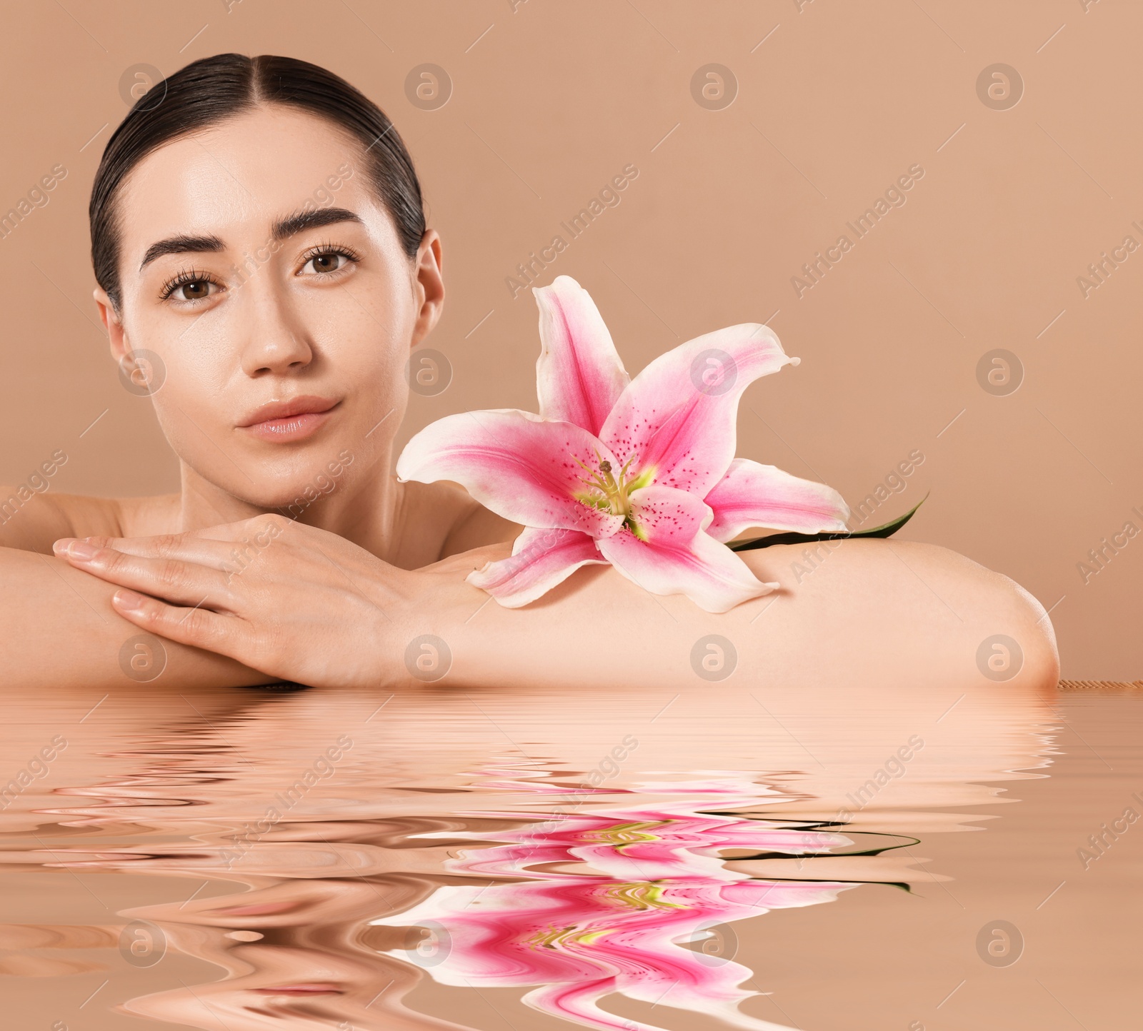 Image of Beautiful woman with perfect skin and lily flower against pale brown background. Her reflection in rippled water