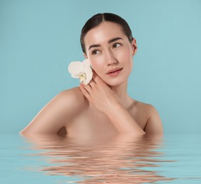 Image of Beautiful woman with perfect skin and orchid flower against light blue background. Her reflection in rippled water