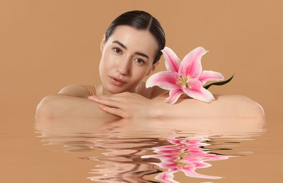 Image of Beautiful woman with perfect skin and lily flower against pale brown background. Her reflection in rippled water